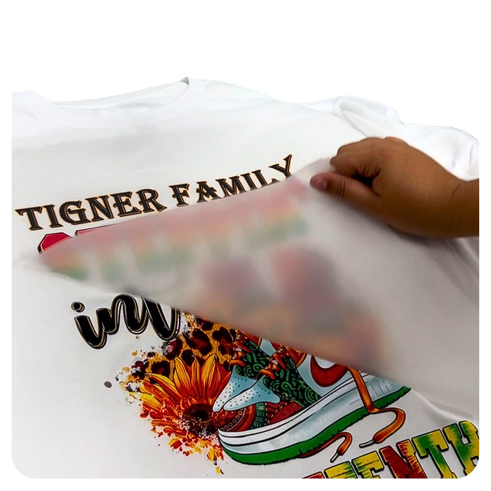 A hand holding a clear plastic sheet with a printed design on it, demonstrating the application of a full-color print on a t-shirt.